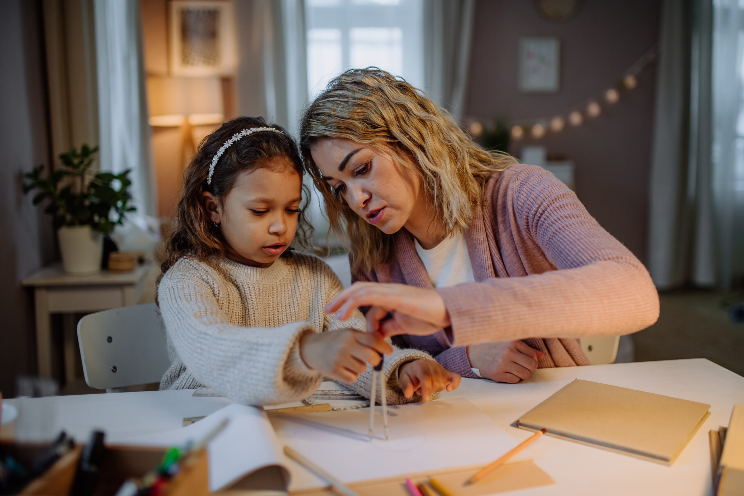 Mother helping her daughter with homework, drawing a circle with comasses in evening at home.
